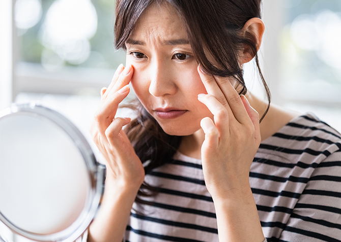 両手で目尻あたりを触りながら鏡を見る女性の画像